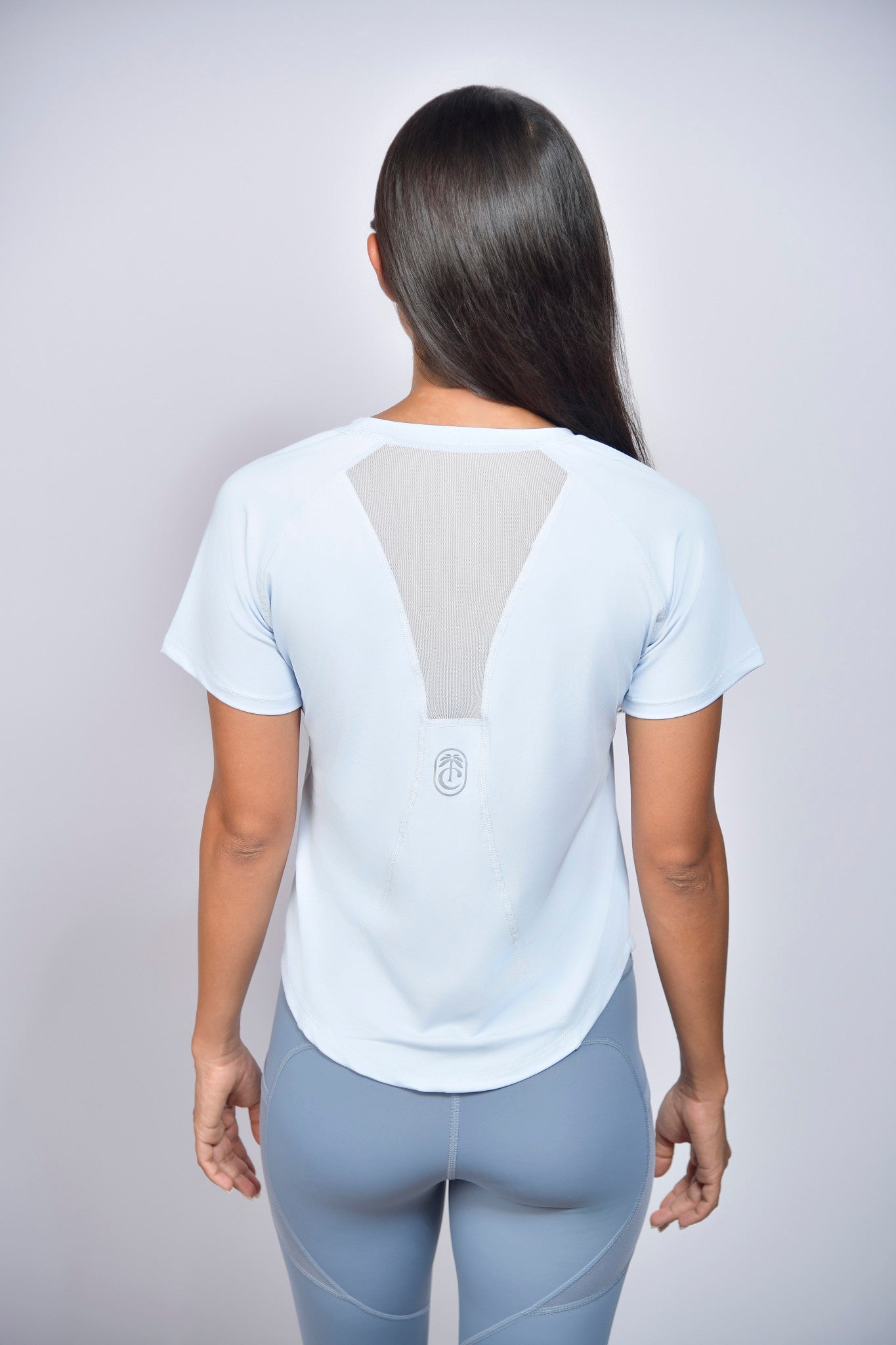 Woman wearing a light-blue t-shirt with  reflective club tropikai on the back against a plain background